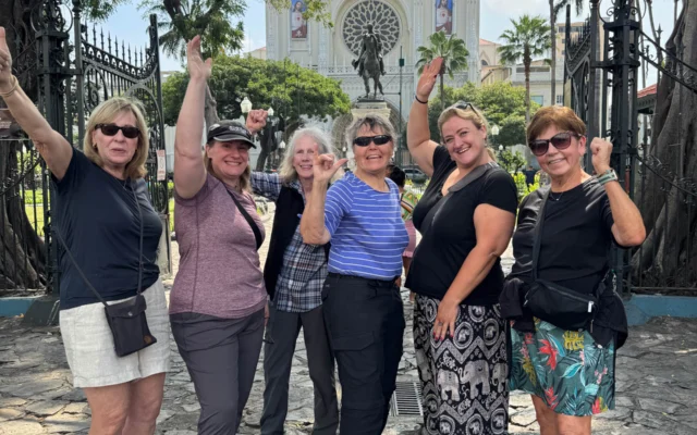 A group of six women pose happily in front of a cathedral with gothic architecture, surrounded by trees and an ornate iron gate.