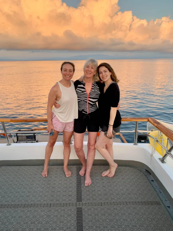Three women standing on a boat deck, smiling with an ocean and cloudy sky in the background.