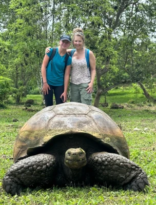 Two people pose behind a large tortoise on grass in a lush, green setting.