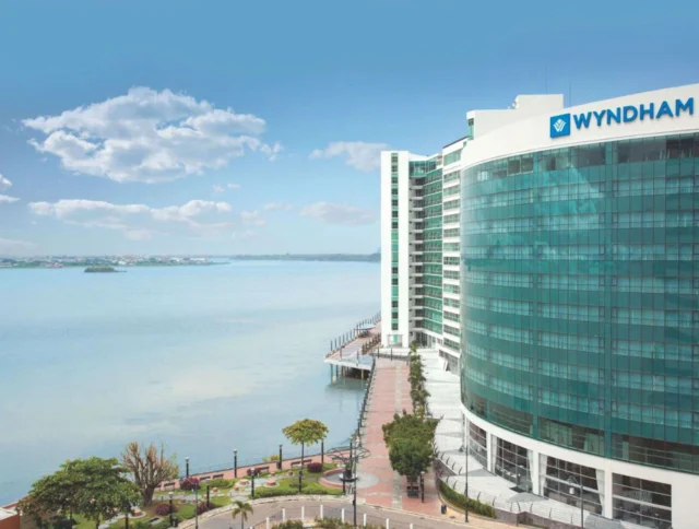 Curved glass-fronted Wyndham hotel beside a calm waterfront, with a paved walkway and distant view of land under a clear sky.