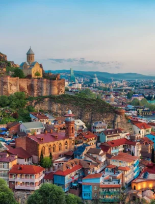 A scenic view of Tbilisi, Georgia, featuring the Narikala Fortress on a hill, surrounded by colorful houses and a sprawling cityscape under a cloudy sky.