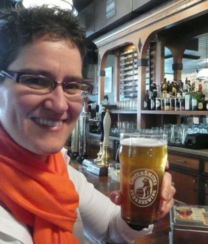 Person holding a glass of beer with a brewery logo, sitting in a bar with various bottles and taps in the background.