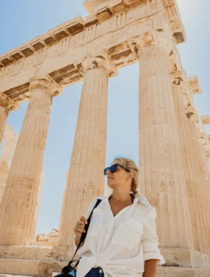 A person in a white shirt and sunglasses stands in front of ancient columns, holding a camera.