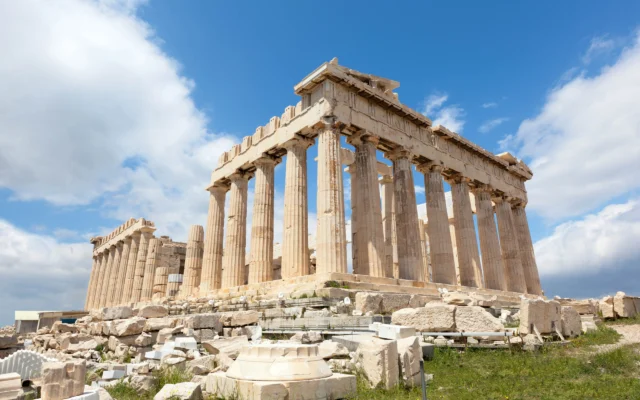 The Parthenon, an ancient temple with Doric columns, sits atop the Acropolis in Athens under a partly cloudy sky. Ruins and scattered stones surround the structure.