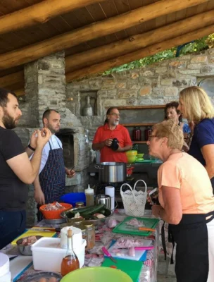 Group of people cooking together in a rustic outdoor kitchen, with ingredients and cookware on the table. Stone walls and wooden roof in the background.