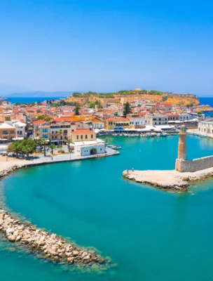 Aerial view of a coastal town with a harbor, surrounded by turquoise water and lined with buildings. A stone pier with a small lighthouse extends into the sea under a clear blue sky.