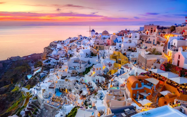 Colorful hillside houses in Santorini, Greece, overlooking the sea at sunset, with a vibrant sky and scattered lights illuminating the buildings.