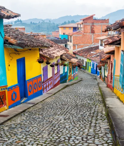 A colorful cobblestone street is lined with vibrant buildings painted in blue, yellow, orange, and green, featuring decorative patterns and wooden shutters, extending down a gentle hill.
