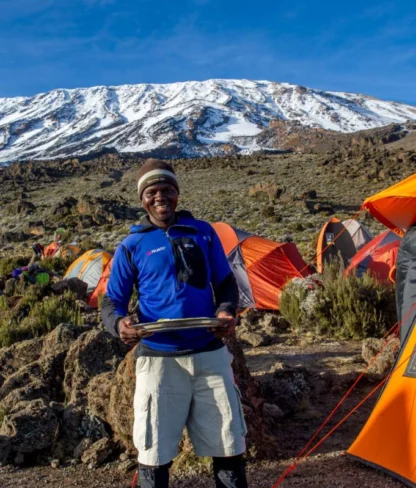 A man stands smiling near a campsite with colorful tents, set against a backdrop of a snow-capped mountain.