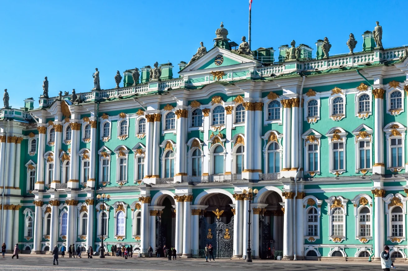 The image shows the Winter Palace in St. Petersburg, Russia, with its ornate green and white facade, columns, and statues under a clear blue sky.
