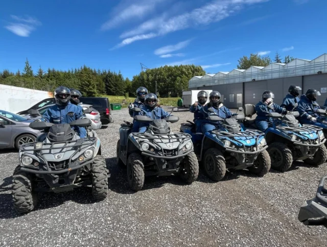 A group of people wearing helmets sit on all-terrain vehicles (ATVs) parked in a row on a gravel surface, with trees and buildings in the background.