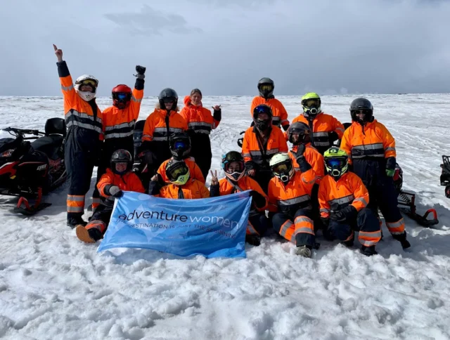 A group of people in orange jumpsuits and helmets pose with an "Adventure Women" banner on a snowy landscape.