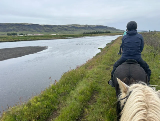 Two people riding horses along a grassy riverbank with a wide view of the landscape, including distant hills under a cloudy sky.