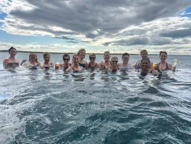 A group of people standing in the ocean, holding drinks, with a cloudy sky in the background.