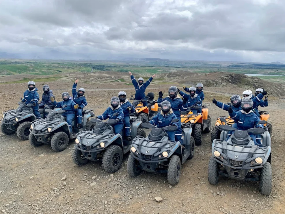 A group of people wearing helmets and blue uniforms sit on all-terrain vehicles, posing with arms raised on a rocky landscape under a cloudy sky.