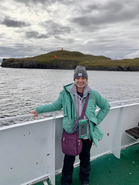 Person in a green jacket and gray beanie stands on a boat deck, smiling with an island and lighthouse in the background under a cloudy sky.