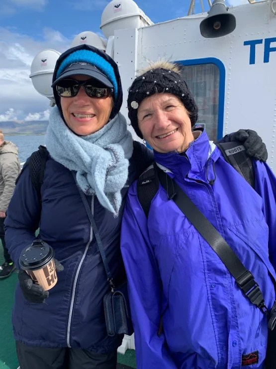 Two smiling people in winter clothing stand together on a boat deck. One holds a coffee cup, and a lifeboat is visible in the background.