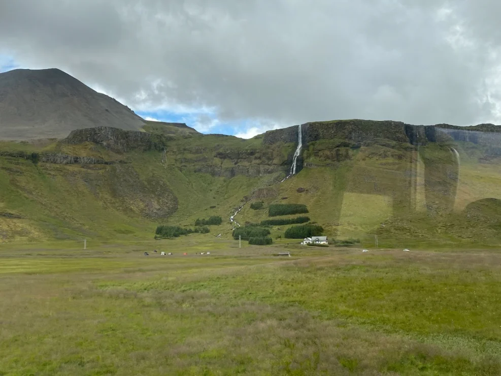 A scenic view of grassy fields, a waterfall cascading down a mountain, and scattered buildings under a cloudy sky.
