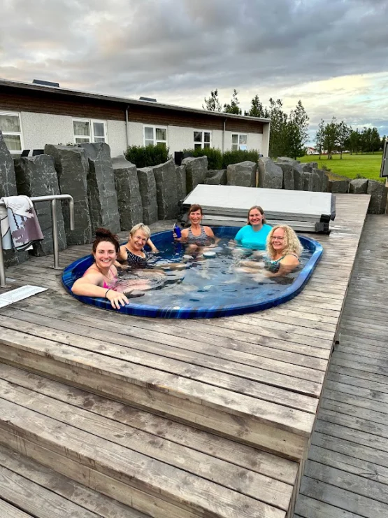 Five people enjoying a hot tub outdoors on a wooden deck, surrounded by stone structures and a building in the background.