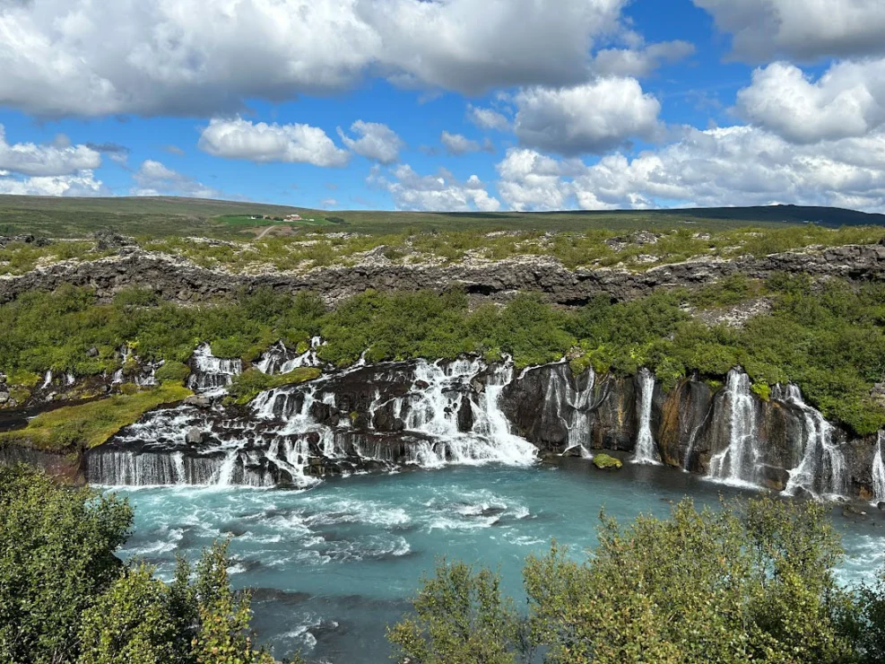 Waterfall with clear blue water cascading over rocky ledges, surrounded by green foliage under a partly cloudy sky.
