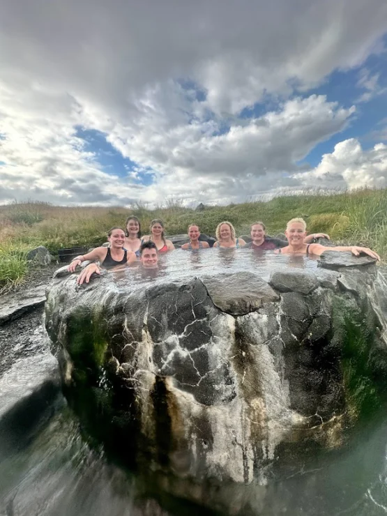 A group of people is sitting in a natural hot spring surrounded by grass under a cloudy sky.