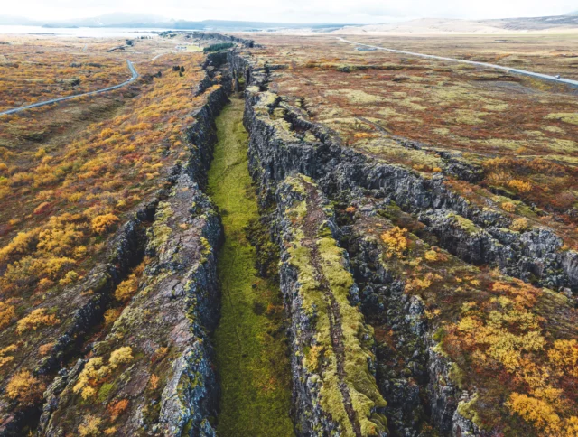 Aerial view of a rocky fissure surrounded by autumn foliage, with a green grassy path running through the middle. Roads and distant hills are visible in the background.