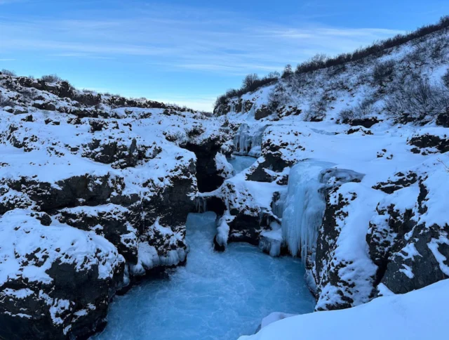 Snow-covered rocky landscape with a partially frozen waterfall and icy river under a clear blue sky.