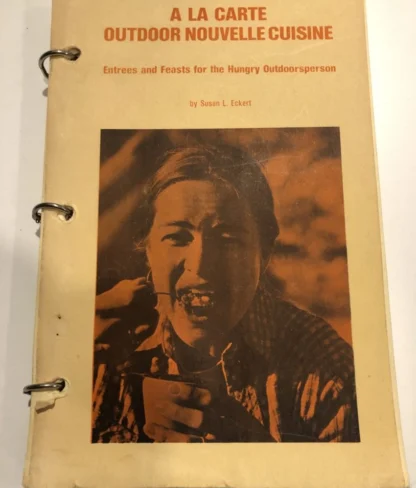 A yellowed cookbook titled "A La Carte Outdoor Nouvelle Cuisine" features a black and white photo of a person eating. The book is bound with metal rings.