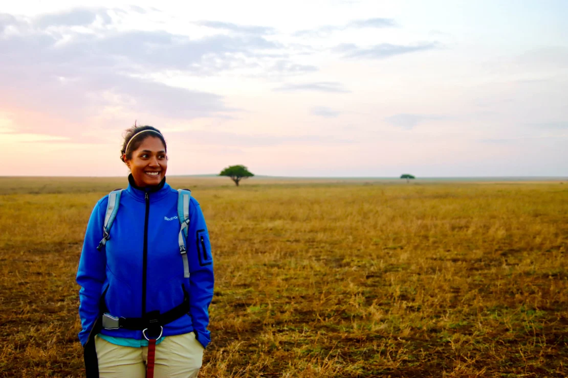 A person in a blue jacket stands in a vast open field at sunset, with distant trees and a cloudy sky in the background.