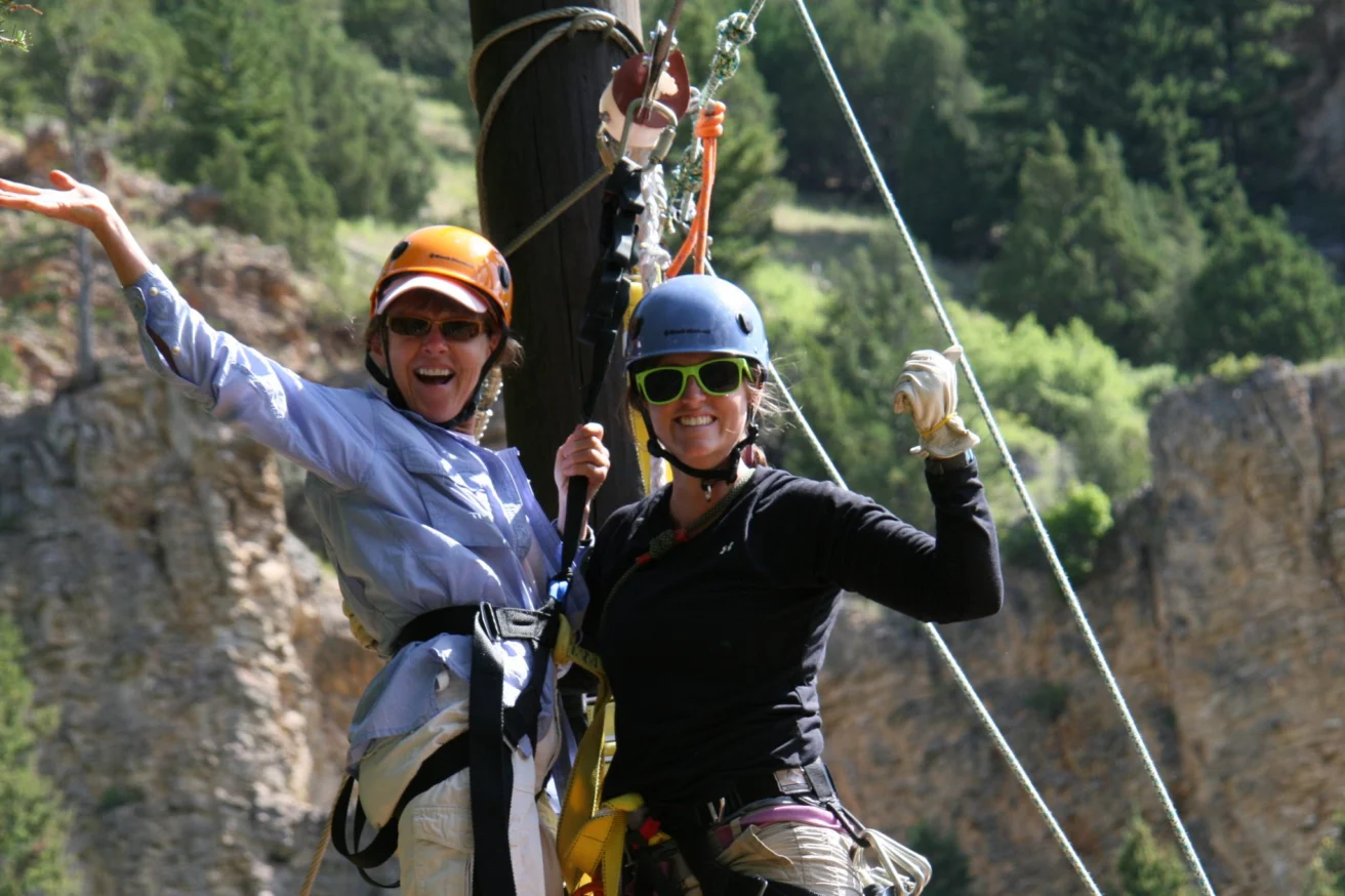 Two people wearing helmets and harnesses are smiling and posing on a zip line against a backdrop of trees and rocky cliffs.