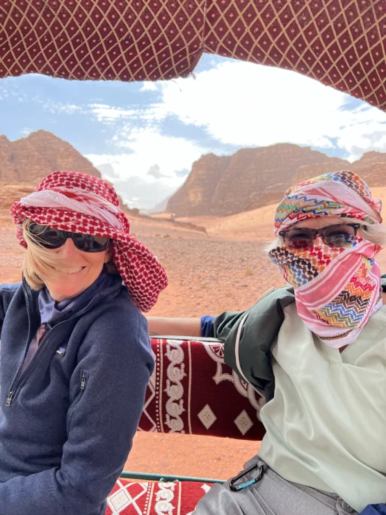 Two people in sunglasses and patterned headscarves sit under a canopy with a desert landscape and hills in the background.
