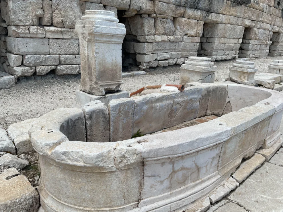 Ancient stone basin with weathered columns and cracked surface, set against a backdrop of stacked stone blocks.