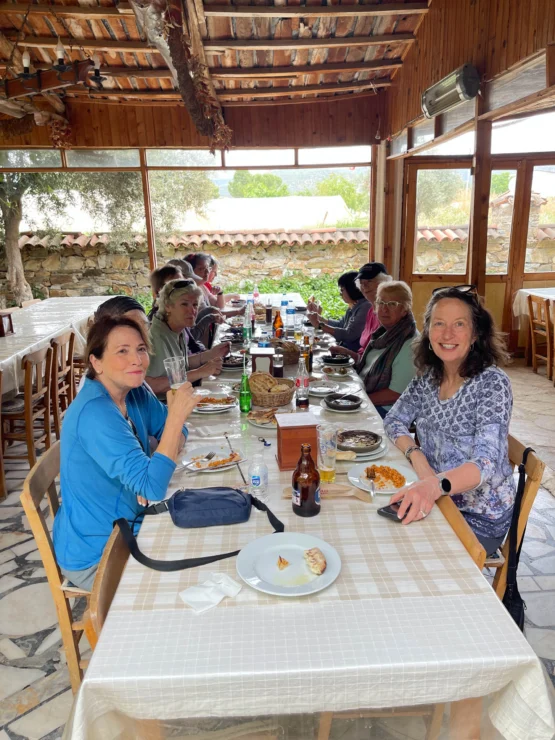Group of people sitting at a long dining table in a rustic restaurant, enjoying a meal with drinks and visible plates of food.