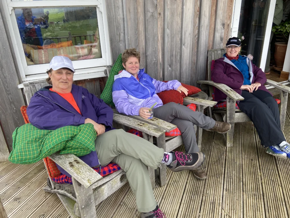Three people sit on wooden chairs outside a rustic building. They are wearing casual outdoor clothing, and two of them have caps. A window and wooden wall are in the background.