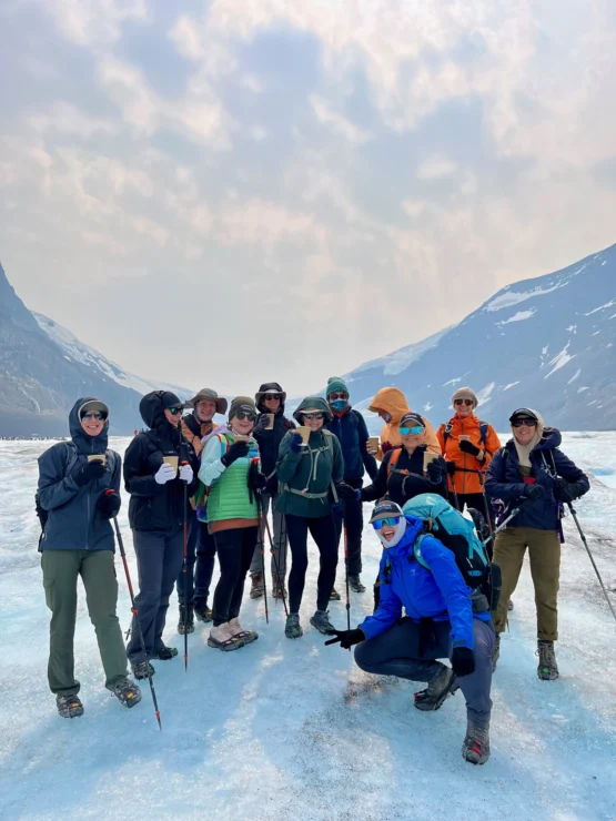 A group of hikers poses on a snowy landscape, equipped with trekking poles and winter gear. Mountains and a cloudy sky are visible in the background.