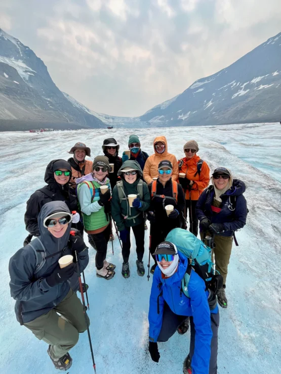 A group of people pose on a glacier, wearing outdoor gear and holding cups. Snow-capped mountains are visible in the background.
