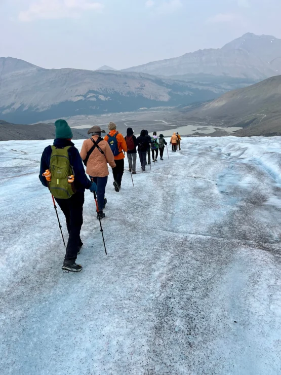 Group of hikers walking in a line on a glacier, surrounded by mountains in the background.