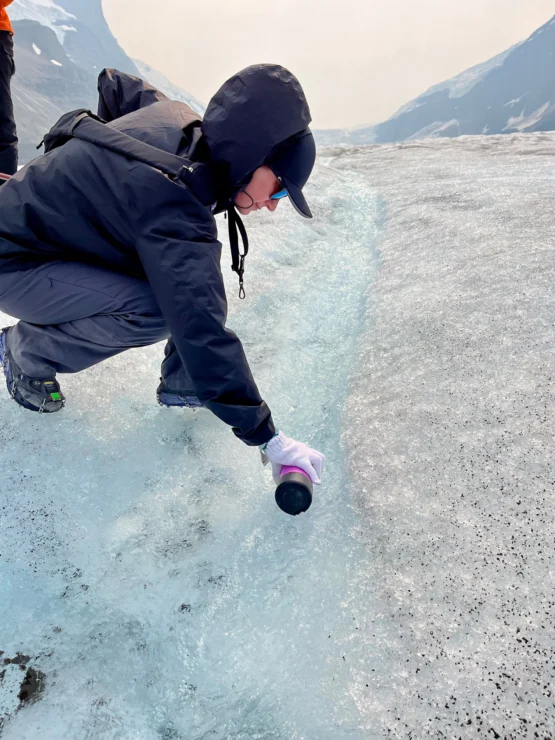 Person in a black jacket and cap collects a sample from an icy surface in a mountainous area.