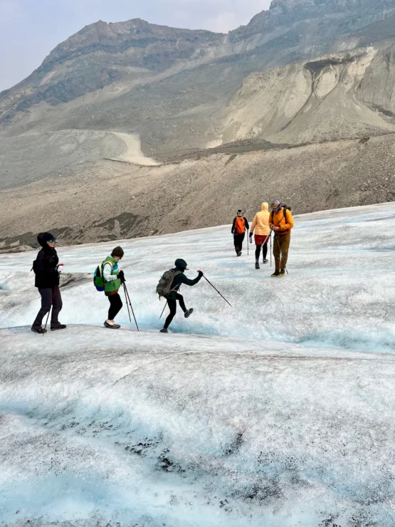 Hikers using trekking poles walk across a glacier with rocky mountains in the background.
