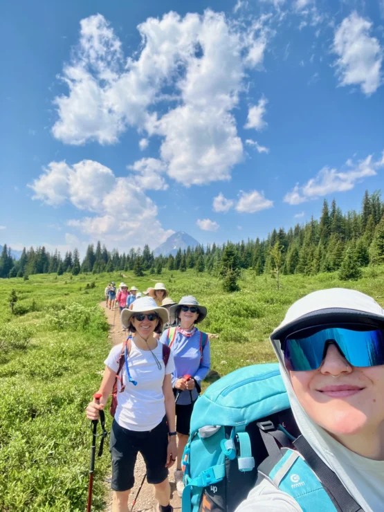 A group of hikers wearing hats and backpacks walks along a trail in a green meadow with a mountain and cloudy sky in the background.