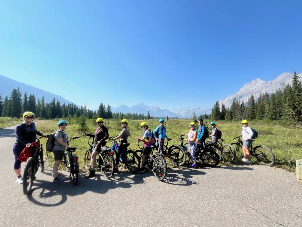 A group of people with bicycles pause on a paved path in a scenic mountain landscape on a sunny day.