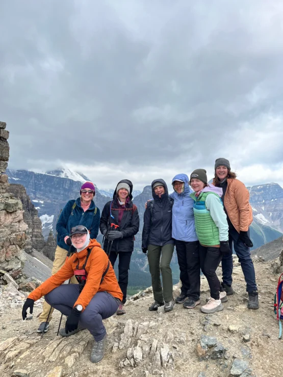 A group of seven people in hiking gear pose together on a rocky mountain summit under a cloudy sky. Snow-capped peaks are visible in the background.
