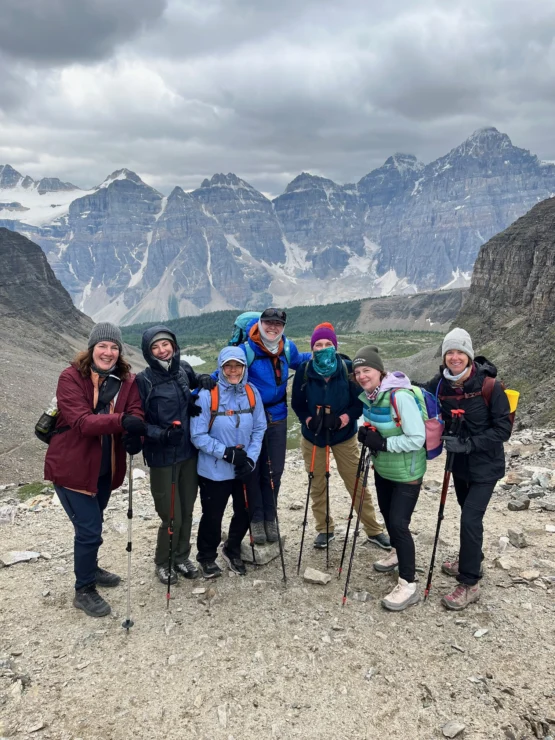 A group of hikers pose together on a rocky terrain with mountain peaks and a cloudy sky in the background.