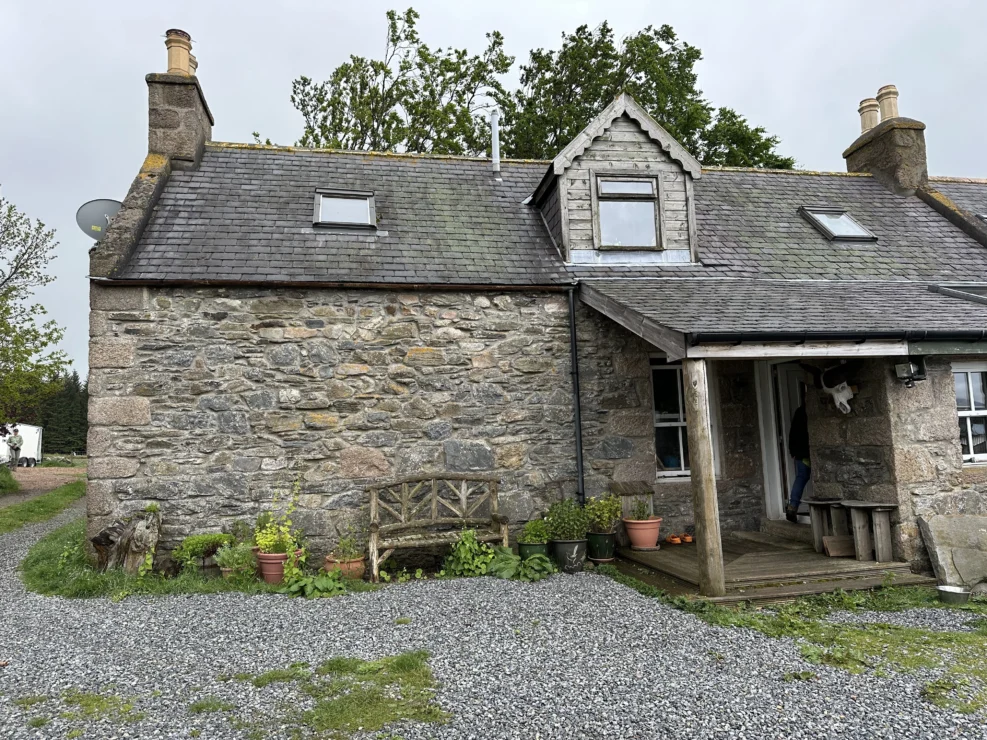 Stone cottage with a slate roof, a small porch, and a bench. Potted plants are arranged near the entrance, and there's a gravel path in front. Trees are visible in the background.