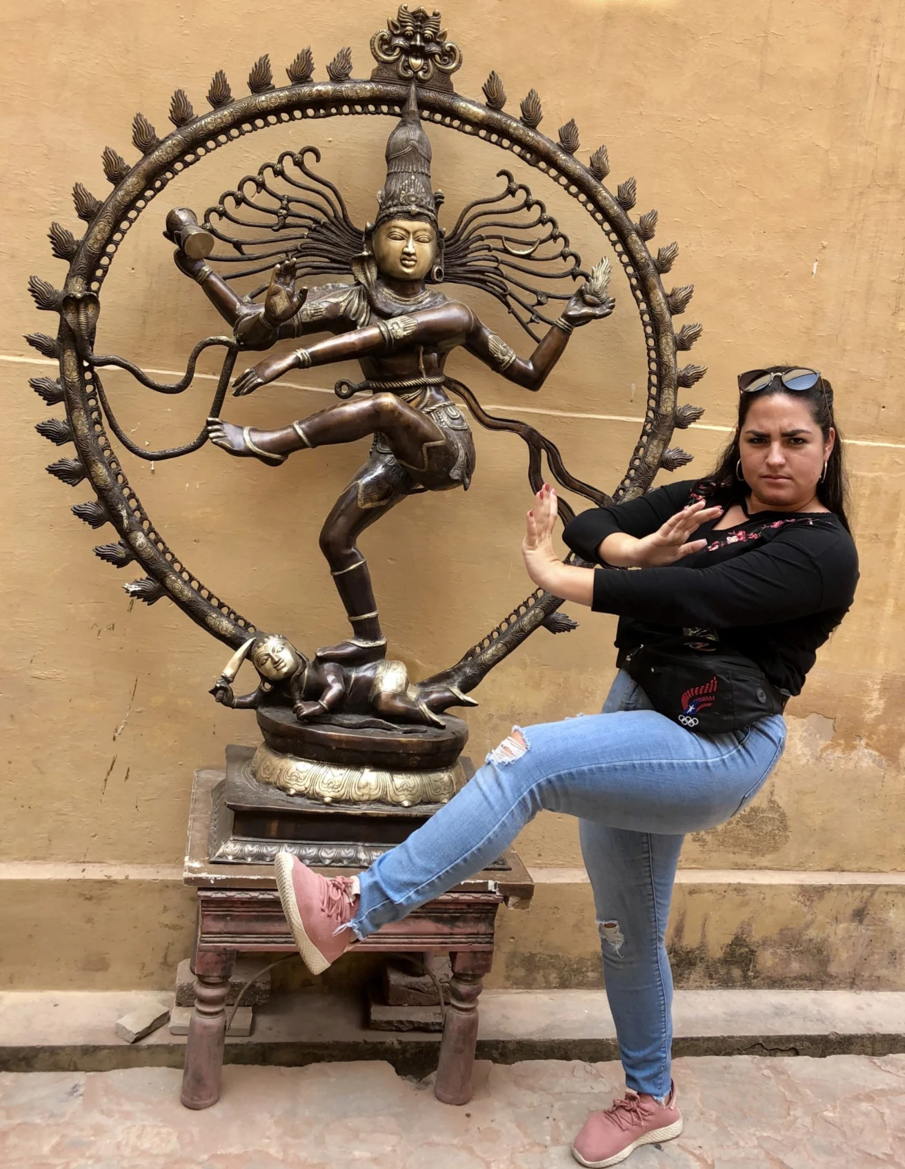 A woman strikes a similar pose to a bronze Nataraja statue, both with raised arms and legs. The statue stands on a wooden pedestal against a beige wall.