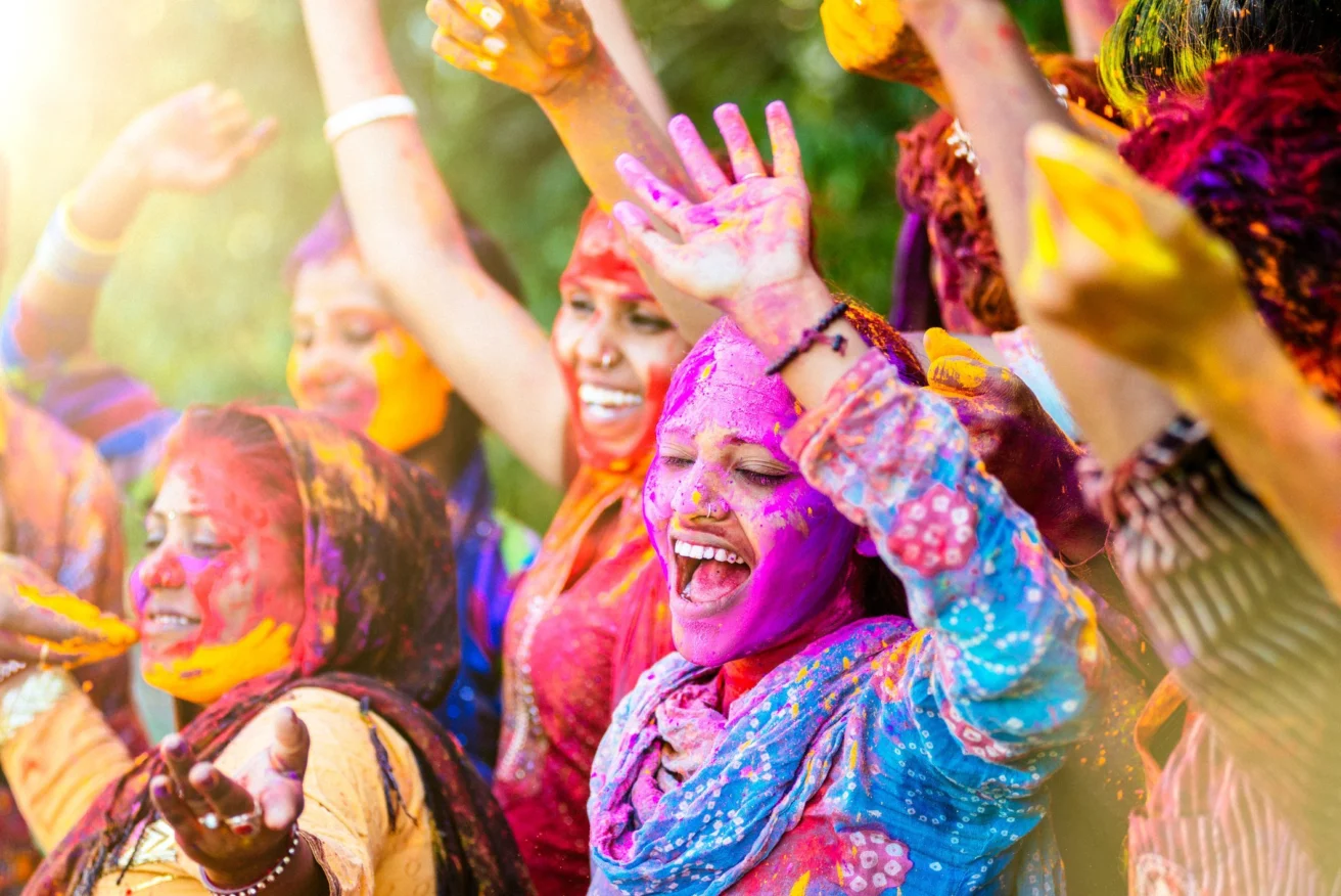 A group of people joyfully celebrating Holi, covered in colorful powders, with arms raised and smiling.
