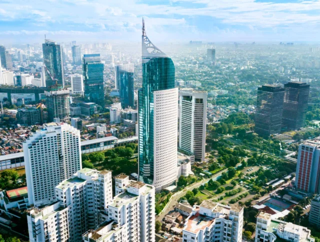 Aerial view of a cityscape with modern skyscrapers, green areas, and scattered residential buildings under a blue sky.