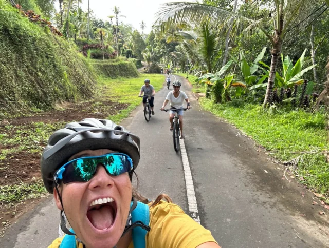 Three people biking on a narrow, lush green road. The person in front is wearing sunglasses and a helmet, smiling widely.