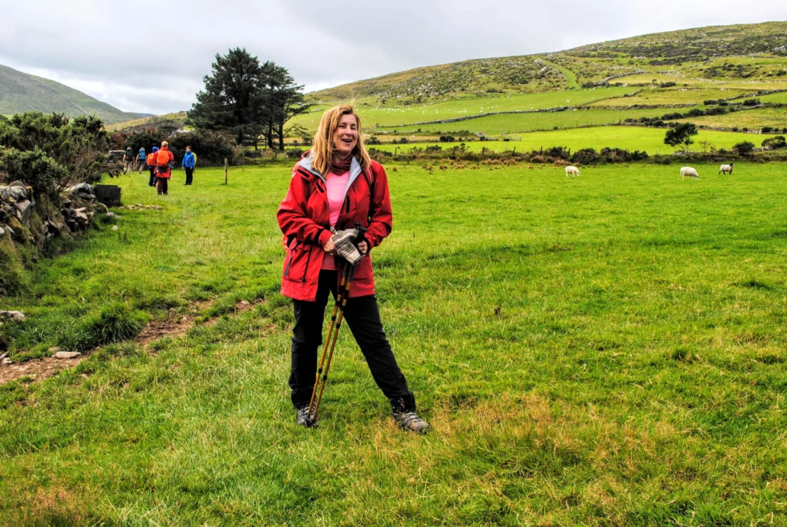 A woman in a red jacket stands with trekking poles on a grassy field, with people and sheep in the background. Hills and trees are visible under a cloudy sky.