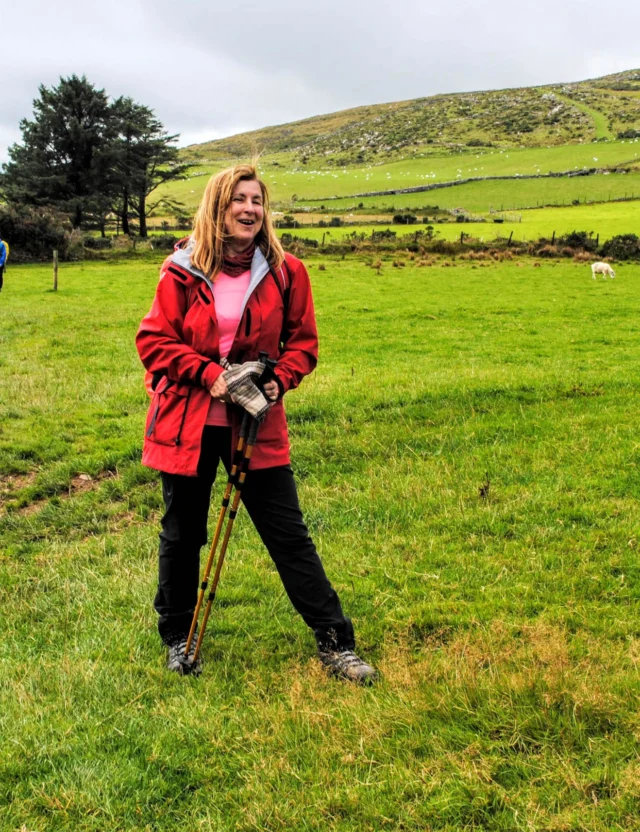 A woman in a red jacket stands with trekking poles on a grassy field, with people and sheep in the background. Hills and trees are visible under a cloudy sky.
